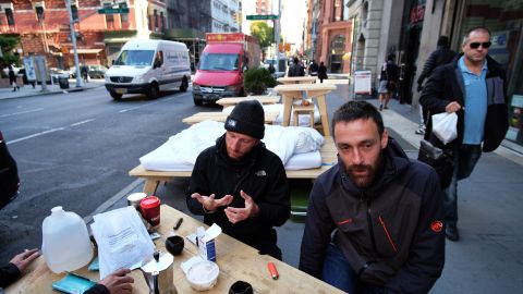 French street artists Sebastien Renauld (R) and Laurent Boijeot sit for their morning coffee next to their beds on a footpath along Broadway in New York on October 20, 2015. The two French street artists are living on the streets of New York for 30 days with their own furniture. On a mission to encourage New Yorkers to slow down, enjoy a cup of coffee, talk to someone new and maybe even take an afternoon nap, the pair make their way down Broadway to the Financial District carrying their white pine tables, chairs, bedding and suitcases, making nearly five blocks each day. AFP PHOTO/JEWEL SAMAD (Photo credit should read JEWEL SAMAD/AFP via Getty Images)