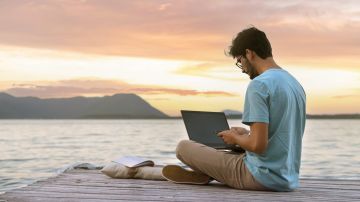 Joven nómada digital sentado en un muelle de madera en el mar trabajando en Internet remotamente al atardecer.