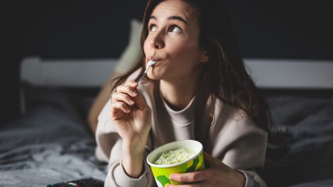 Mujer disfruta de un bote de helado, recostada en su cama.