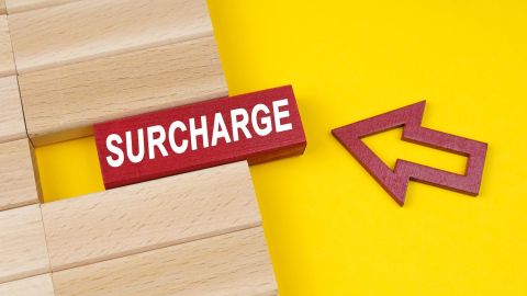 Sobre una superficie amarilla hay bloques de madera y una flecha que apunta a un bloque con la inscripción - Surcharge (Recargo).