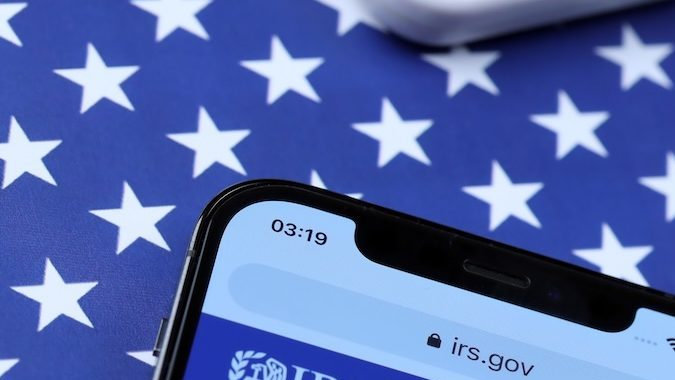 Sitio web oficial del IRS en la pantalla del iPhone 12 pro con formularios de impuestos en papel 1040 y w-2 en la bandera de los Estados Unidos.