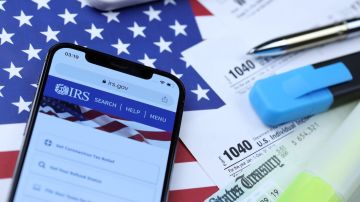 Sitio web oficial del IRS en la pantalla del iPhone 12 pro con formularios de impuestos en papel 1040 y w-2 en la bandera de los Estados Unidos.