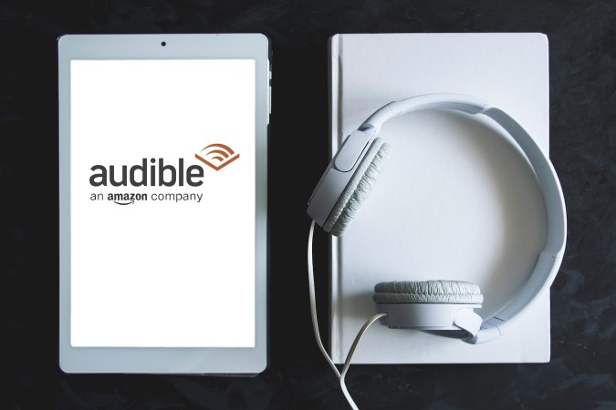 Logotipo de la pantalla de la aplicación Audible en una tableta, junto a unos auriculares blancos.