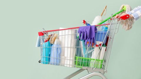 Mujer con un carrito de la compra lleno de productos de limpieza sobre un fondo verde pálido.
