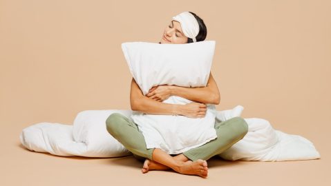 Mujer latina joven, tranquila y sonriente, de cuerpo entero, viste pijama, duerme con antifaz, descansa y se relaja en casa sentada con una almohada.