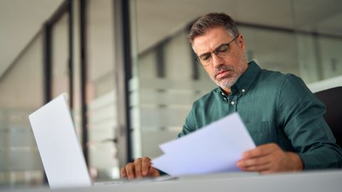 Ejecutivo de mediana edad ocupado, director ejecutivo maduro, gerente de cuentas de recursos humanos masculino sosteniendo documentos usando una computadora portátil en la oficina, verificando la factura financiera, los datos en el informe, haciendo una descripción general del plan de archivo de papel contable.