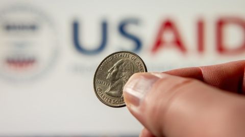 Moneda de 25 centavos de dólar estadounidense sostenida con los dedos sobre un fondo con el logotipo de USAID.