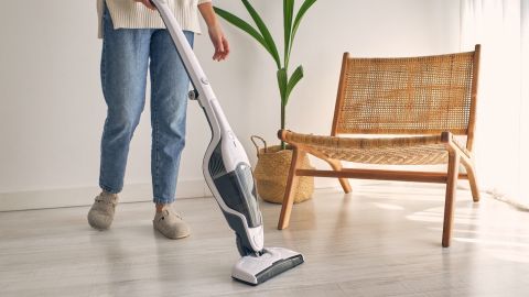 Vista lateral de ama de casa sin rostro con atuendo casual y pantuflas limpiando piso laminado con aspiradora vertical moderna en un apartamento minimalista y luminoso.