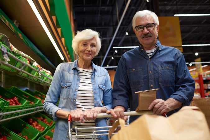 Retrato de una pareja mayor moderna con carrito de compras posando y mirando a la cámara en el supermercado mientras disfruta de las compras.