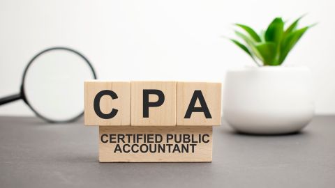 Cubos de madera con letras CPA Contador Público Certificado sobre mesa blanca con teclado y gafas