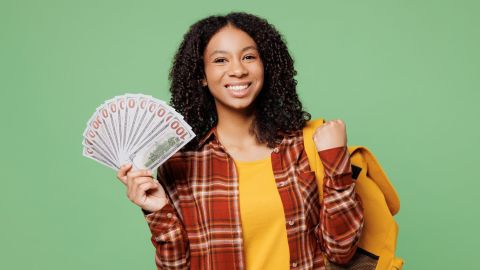 Una joven estudiante lleva una mochila con ropa informal de color marrón y amarillo y sostiene un abanico lleno de billetes de dólar en forma de ganadora aislada sobre un fondo verde liso. Concepto de escuela secundaria y universidad.